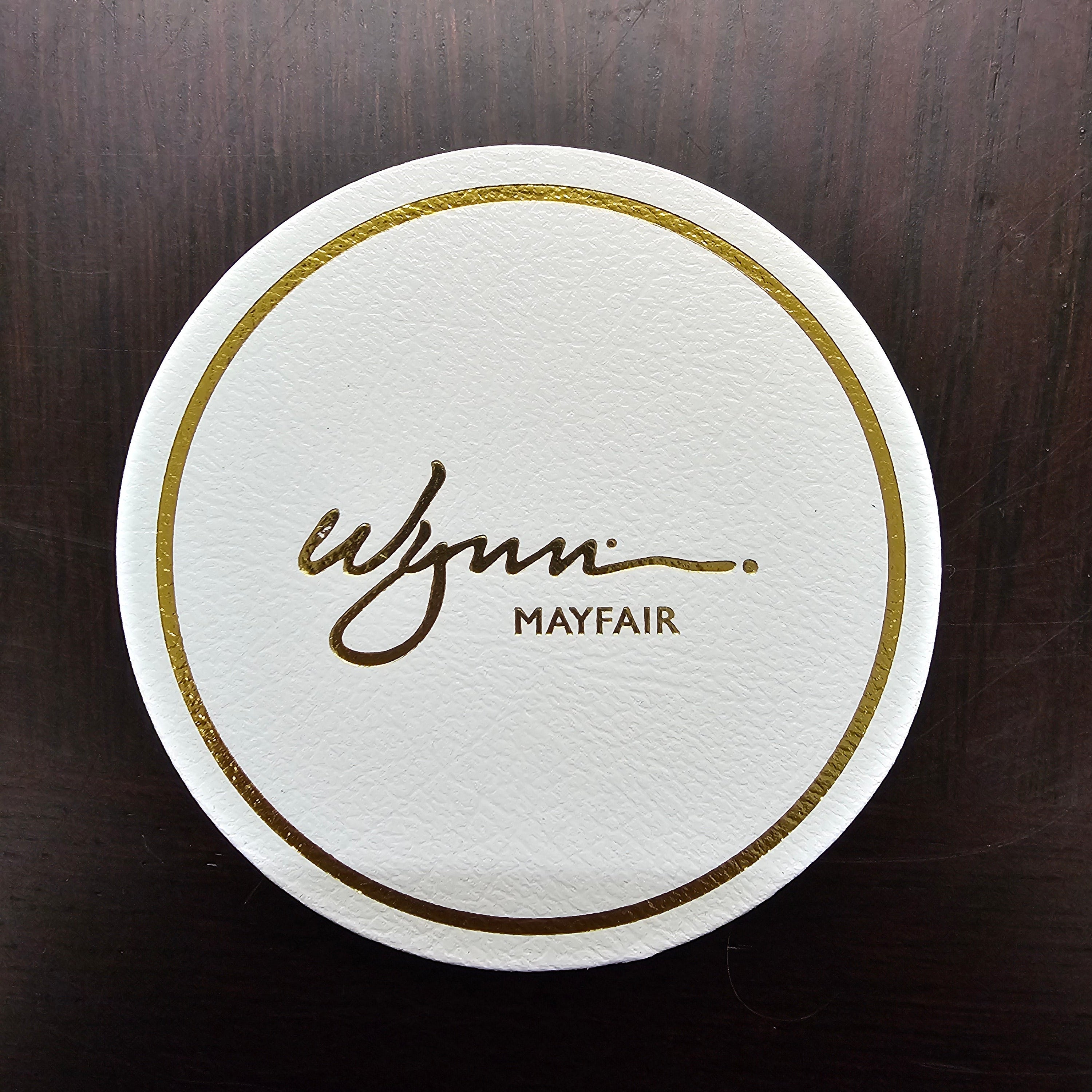 Round white coaster with gold border and 'Mayfair' branding on a dark wooden surface
